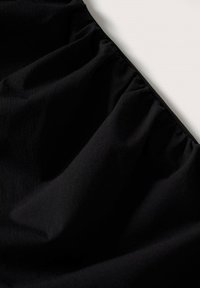 Plis de tissu noir rassemblés visibles sur une surface claire unie, montrant la texture et la draperie douce du matériau.