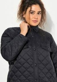 Chaqueta negra acolchada con cuello, cierre frontal de botones y dos bolsillos en el pecho. La tela parece suave y brillante, proporcionando aislamiento.