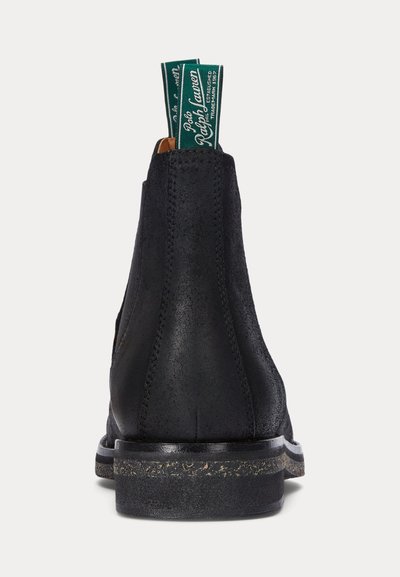 Bota de cano curto em pele preta com textura lisa, sola de cor clara contrastante e alças proeminentes no calcanhar com marcação verde.