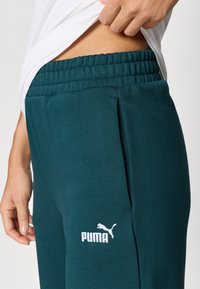 Teal sweatpants with an elastic waistband, side pocket, and white Puma logo on the left thigh. Soft material with a smooth texture.