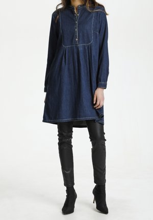 Vrouw draagt een donkerblauwe denim tuniekjurk met lange mouwen over zwarte leren broek en zwarte enkellaarsjes met hoge hakken, staande tegen een witte achtergrond.