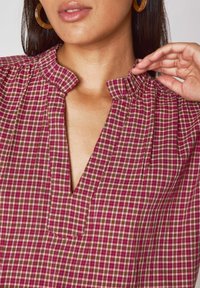 Blusa a quadri rossi e verdi con scollo a V, dettagli arricciati sulle spalle e tessuto di cotone testurizzato.