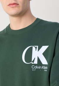 Sweatshirt din bumbac verde închis cu guler rotund și nervuri. Prezintă un logo alb proeminent, "CK Calvin Klein EST - 1968," pe față.