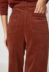 Pantaloni in velluto a coste marroni con una vestibilità a gamba larga. Presentano due tasche posteriori e un tessuto testurizzato con coste visibili.