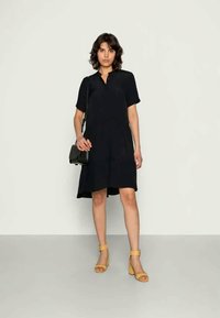 Vestido preto de mangas curtas com placket frontal, comprimento até ao joelho e um corte relaxado, acompanhado por sandálias de salto amarelas e uma pequena mala preta.