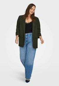 Sieviete staigā pārliecinoši, valkājot olīvu zaļa krāsas žaketi, melnu kamzolīti ar mežģīņu apdari, gaismas zila augsto vidukli džinsus un tumšas, smailas kurpes.