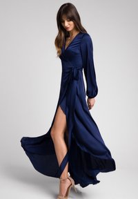 Granatowa sukienka typu wrap z satynowego materiału z długimi rękawami, wysokim rozcięciem oraz wiązaniem w talii, z falującą spódnicą i gładką teksturą.