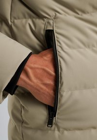 Doudoune beige avec une poche zippée sur le côté. Le tissu semble lisse avec des sections matelassées. Un poignet noir est visible.