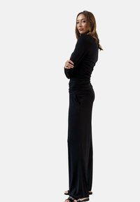 Czarna sukienka z długim rękawem o marszczonym wykończeniu w talii, z dopasowanym górnym fasonem i szerokimi nogawkami. Model stoi z założonymi rękami.