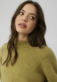 Pull en maille vert olive, avec un col montant et des manches raglan. Texture douce avec une coupe légèrement ample et un motif de tricot visible.