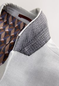 Nahaufnahme eines hellgrauen Jackenkragens mit schwarzer geometrischer Naht und gemustertem braun-blau-beigem Innenfutter.