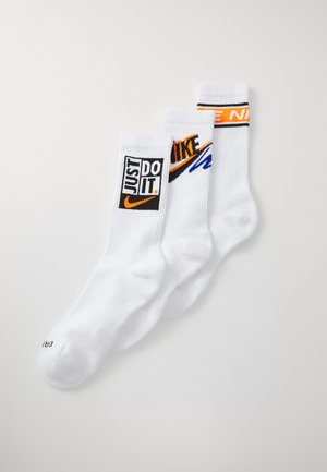 Tři páry bílých ponožek s kontrastními černými a oranžovými logy: "JUST DO IT," "NIKE," a proužkované akcenty. Rýhovaná textura.