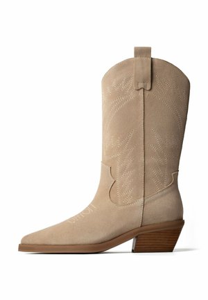 Bottine beige en daim style western avec bout pointu, surpiqûres décoratives, languette de traction et talon bloc empilé en bois.