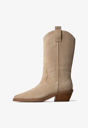 Beżowy zamszowy botek w stylu western z ostrym noskiem, ozdobnym przeszyciem, pętelką do ściągania i piętą typu stacked z drewna.