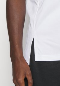 Maglietta atletica bianca con un tessuto testurizzato, aperture laterali e una vestibilità rilassata, abbinata a pantaloni corti neri atletici. Mano posizionata sulla maglietta.