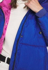 Puffy jacket in blue and pink colors, made of shiny material. Features a zipper closure, button accents, and quilted texture.