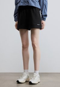 Μαύρο βαμβακερό σορτς με ελαστική ζώνη μέσης και μικρό λογότυπο μάρκας στο στρίφωμα. Φορεμένο με λευκές αθλητικές κάλτσες και ανοιχτόχρωμα αθλητικά παπούτσια.