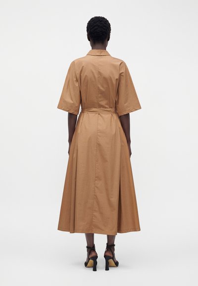 Femme debout, de dos, portant une robe midi couleur sable avec des manches aux coudes, une ceinture et des sandales noires à talons ouverts au niveau des orteils.
