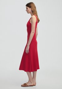Červené midi šaty bez rukávů se fitted vrškem a rozšířenou sukní, vyrobené z žebrované látky. Modelka má na sobě ploché sandály s otevřenou špičkou.