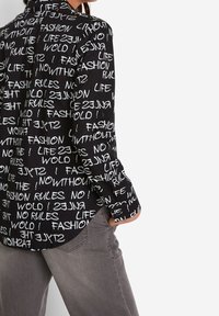 Chemise noire à boutons avec des motifs de texte blancs portant les mots "MODE", "VIE" et "RÈGLES". Manches longues avec accents de boutons, coupe décontractée.