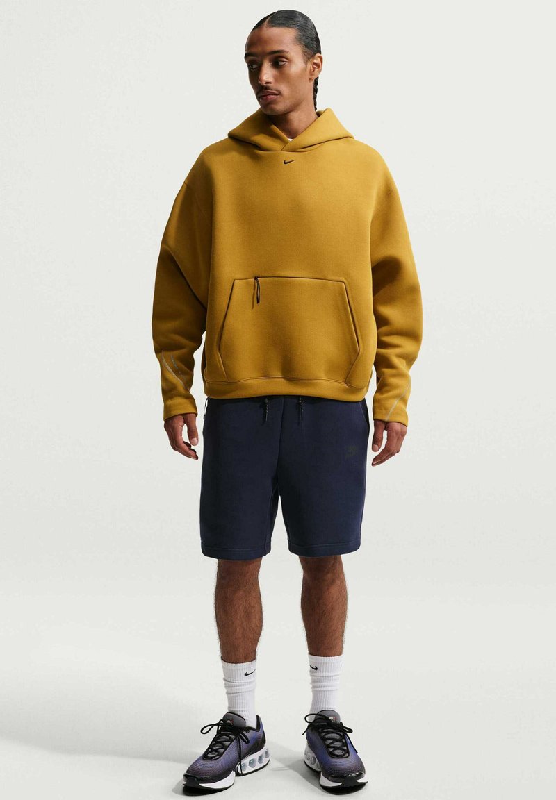 Man staand in een mosterdgele hoodie, marineblauwe korte broek, witte sokken en paarse sneakers tegen een effen lichte achtergrond.