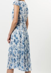 Robe midi plissée blanche avec motif floral bleu. Caractérisée par des manches courtes, une taille cintrée, et un design fluide pour faciliter le mouvement.