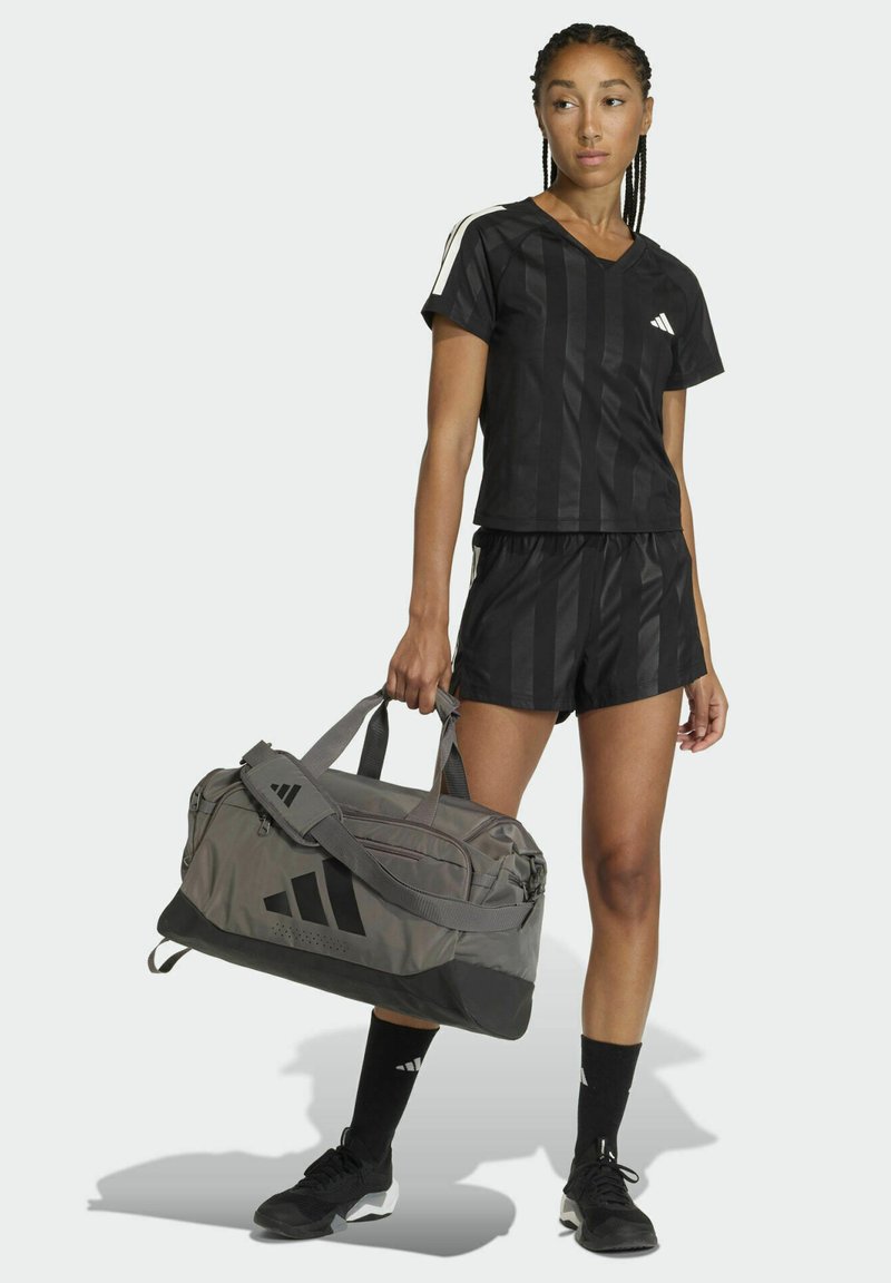 Camiseta deportiva negra y pantalones cortos con rayas verticales, combinados con una bolsa de viaje gris con el logo. Calcetines negros y zapatos deportivos completan el conjunto.