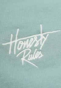 Ljust mintgrön tyg med vit broderad text som lyder "Ärlighet Regler," med en mjuk textur och minimalistisk design.