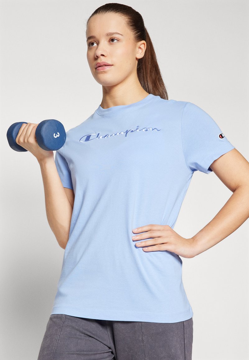 T-shirt azzurro chiaro con logo "Champion" in corsivo, maniche corte e scollatura tonda, abbinato a un manubrio blu scuro contrassegnato da '3'.