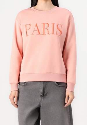 Femme portant un sweat-shirt rose avec "PARIS" brodé à l'avant et un jean gris ample, visible du cou jusqu’aux hanches.
