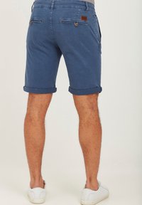 Blue rolled-up shorts made of cotton, featuring two back pockets with buttons, and a brown leather accent near the waistband. White shoes visible.