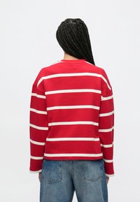 Chemise à manches longues rayée rouge avec des rayures horizontales crème, encolure ronde et coupe décontractée. Le matériau semble doux et légèrement texturé.