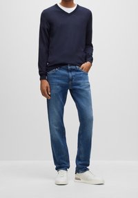 Marinblå v-hals tröja, denimblå raka jeans och vita sneakers. Tröjan har åtsittande ärmslut; jeansen har traditionella fickor och knappgylf.