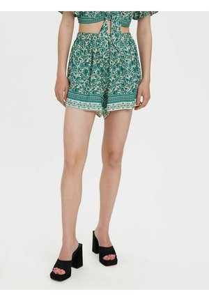 Frau trägt grüne, hoch taillierte Shorts mit Blumenmuster, kombiniert mit schwarzen Sandalen mit offenem Zehenbereich und Blockabsatz vor weißem Hintergrund.
