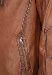 Bruine leren jas met een gestructureerde afwerking, opvallende stiksels en een zijzak met rits en zilverkleurige hardware. Gladde oppervlakte en getailleerde vorm.