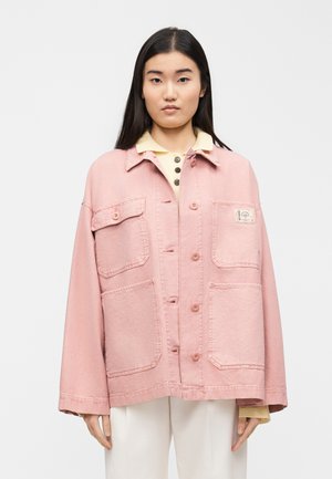Femme aux longs cheveux noirs portant une veste-chemise rose surdimensionnée et une chemise de couleur crème, debout devant un fond blanc uni.