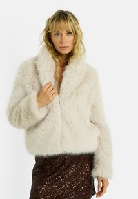 Veste en fausse fourrure beige clair avec un large col et de longues manches duveteuses. Le matériau texturé ajoute du volume, associé à une jupe brune à sequins.