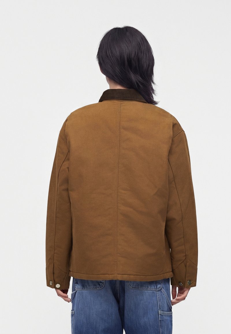 Person with dark hair wears a brown jacket with snap button cuffs and blue denim jeans, facing away against a plain light background.