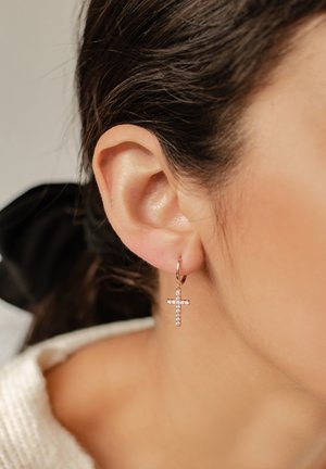 Boucles d'oreilles créoles en ton or avec un pendentif en forme de croix orné de strass transparents, suspendues à un petit fermoir. Vue rapprochée de l'oreille.