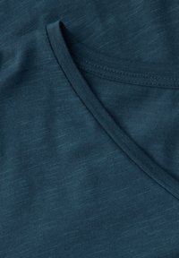 Smaragdově zelená pletenina s jemnou texturou. Obsahuje límec ve tvaru V a hladký povrch. Materiál je lehký a mírně elastický.