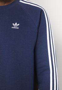 Tamno plavi dukserica s bijelim prugama na ramenima, s malim bijelim Adidas logotipom na prsima. Pamuk, teksturirana površina.