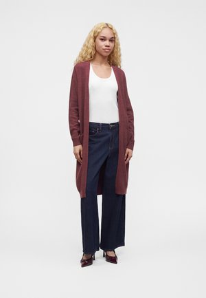 Femme aux cheveux blonds bouclés portant un long cardigan bordeaux, un débardeur blanc, un jean large foncé et des talons bordeaux, debout devant un fond blanc.