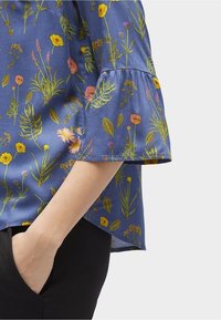 Blouse fleurie bleue présentant un mélange de motifs jaunes, roses et verts, avec un détail de manche à volants et une coupe décontractée.