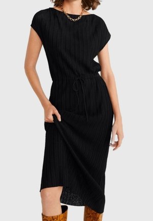 Frau trägt ein schwarzes, geripptes Midikleid mit kurzen Ärmeln und einem Tunnelzug in der Taille, kombiniert mit braunen Stiefeln mit Schlangenhautmuster.
