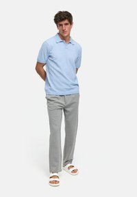 Lyseblå kortermet polo med teksturert stoff, kombinert med lysegrå bukser og hvite sandaler, står med hendene bak ryggen.