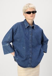 Denim liela izmēra krekls ar sirdspapīrveida izgriezumiem, pogu aizdari priekšā un plašām piedurknēm. Krāsa ir tumši zila ar izturīgu tekstūru.