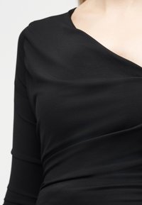 Γυναίκα που φοράει εφαρμοστή μαύρη μπλούζα με φαρδύ ντεκολτέ και λεπτομέρειες με σούρες στο στήθος και τη μέση.