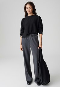 Schwarzer Pullover mit strukturierter Oberfläche und dreiviertel Ärmeln, kombiniert mit grauen Hose mit weitem Bein. Die Person hält eine schwarze Tasche. Schwarze Schuhe vervollständigen das Outfit.