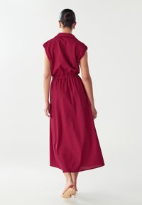 Donna con i capelli raccolti in un chignon, che indossa un vestito midi senza maniche di color borgogna con vita arricciata e mule beige con tacco alto, di spalle.