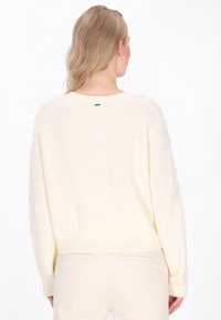 Crème-kleurige gebreide trui met een ribbelstructuur, ronde halslijn en lange mouwen, voorzien van een subtiele logo-label op de achterkant.